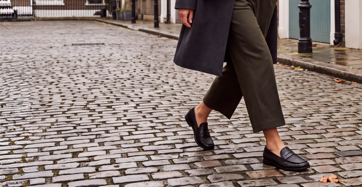 Clover Woven Loafer in Black hand-woven leather, worn mid-stride on wet cobbles with olive cropped trousers and a dark coat
